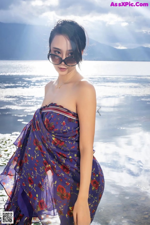 A woman in a purple dress and sunglasses posing by the water.