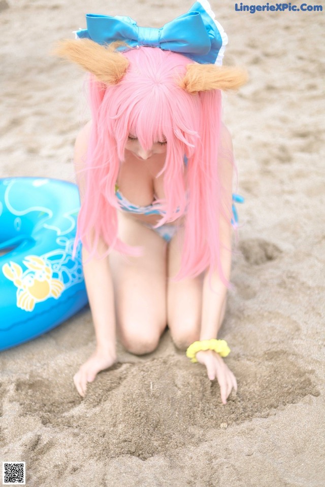 A woman with pink hair and a blue bow sitting in the sand.