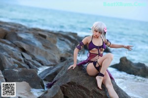 A woman in a purple bikini sitting on a rock by the water.